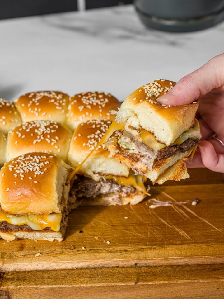 A wooden board with cheeseburger sliders with a hand pulling a slider from the set. The board is on a marble counter.