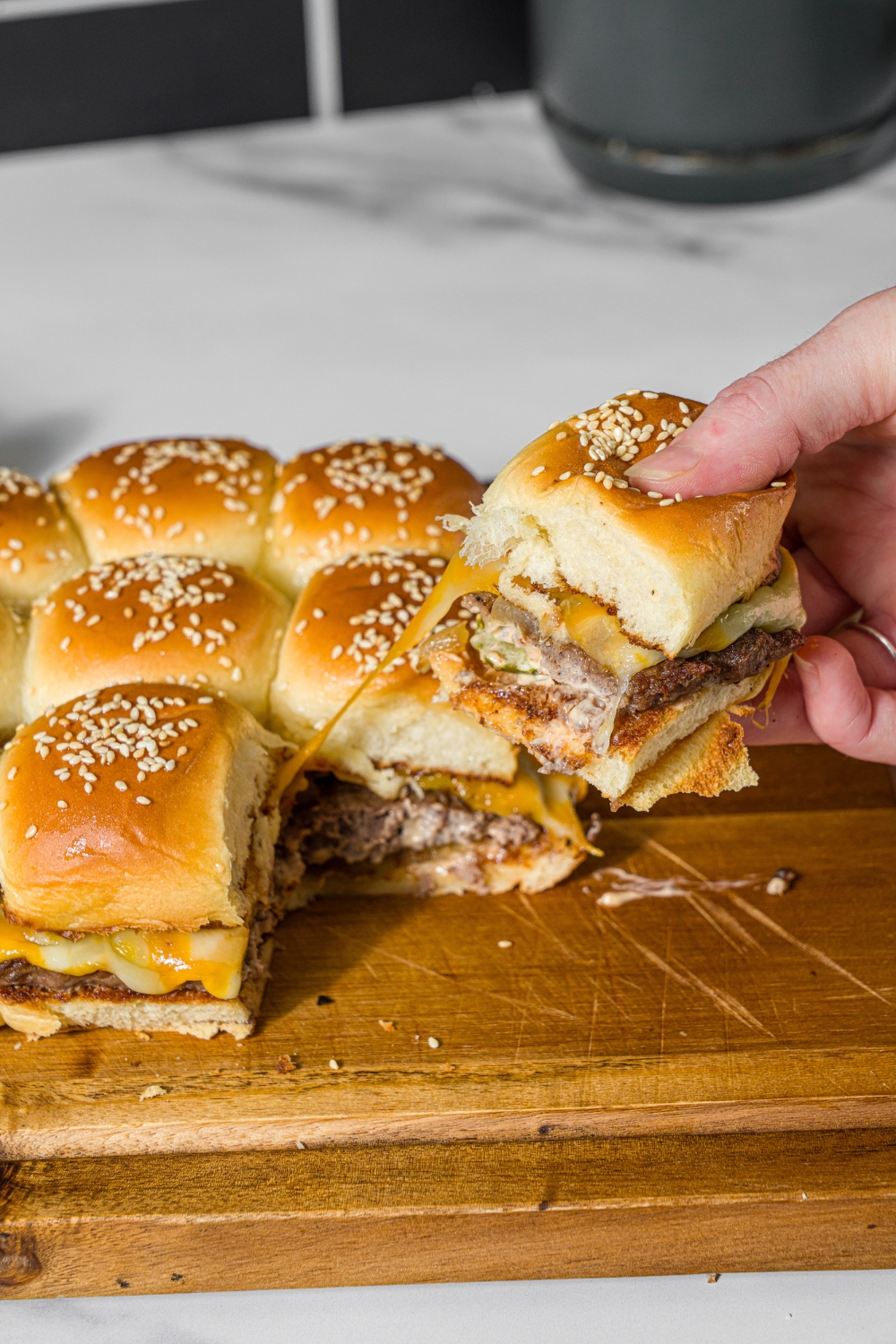 A wooden board with cheeseburger sliders with a hand pulling a slider from the set. The board is on a marble counter.