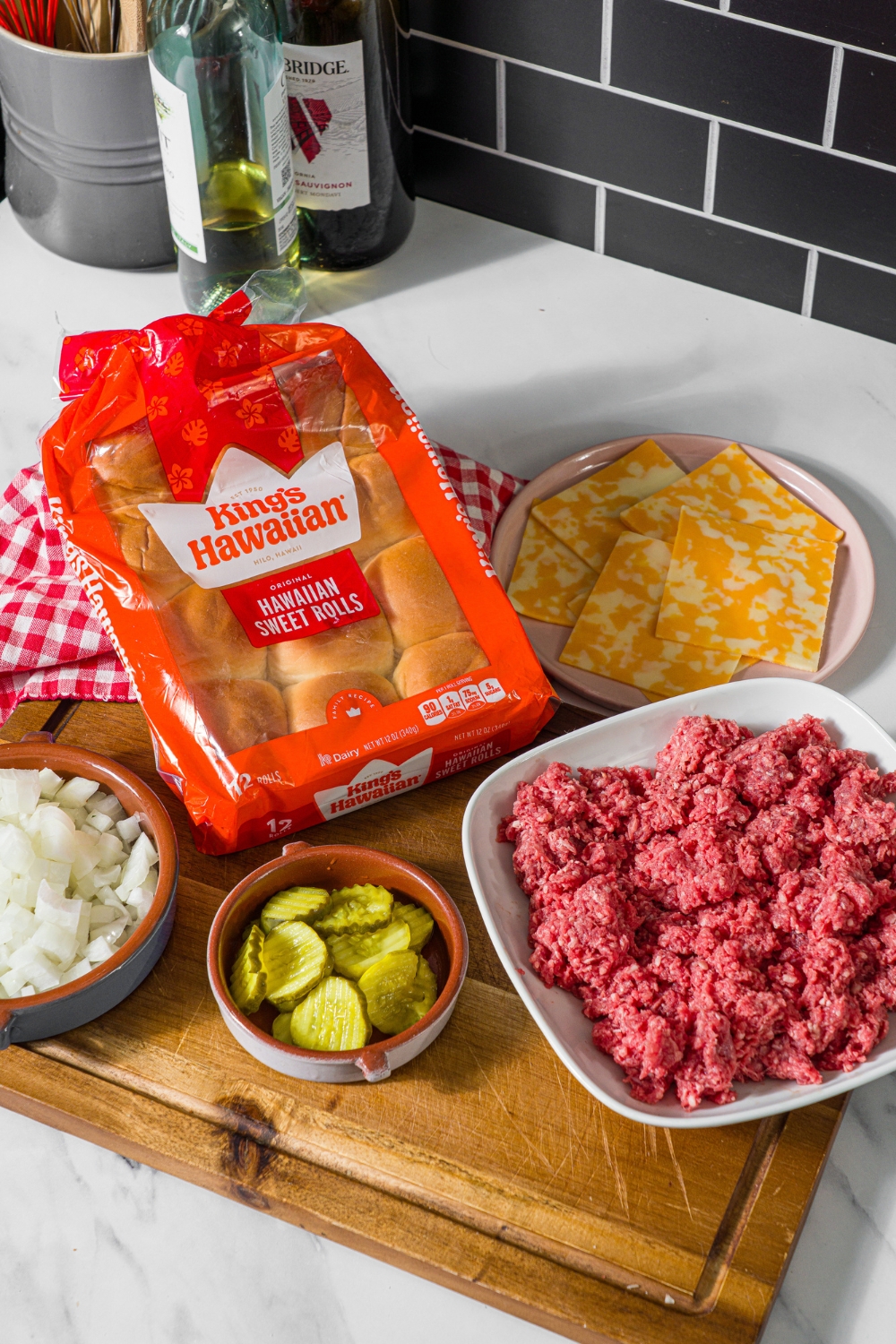 A wooden board with ingredients to make cheeseburger sliders including hawaiian rolls, ground beef, pickles, onion, and cheese slices.