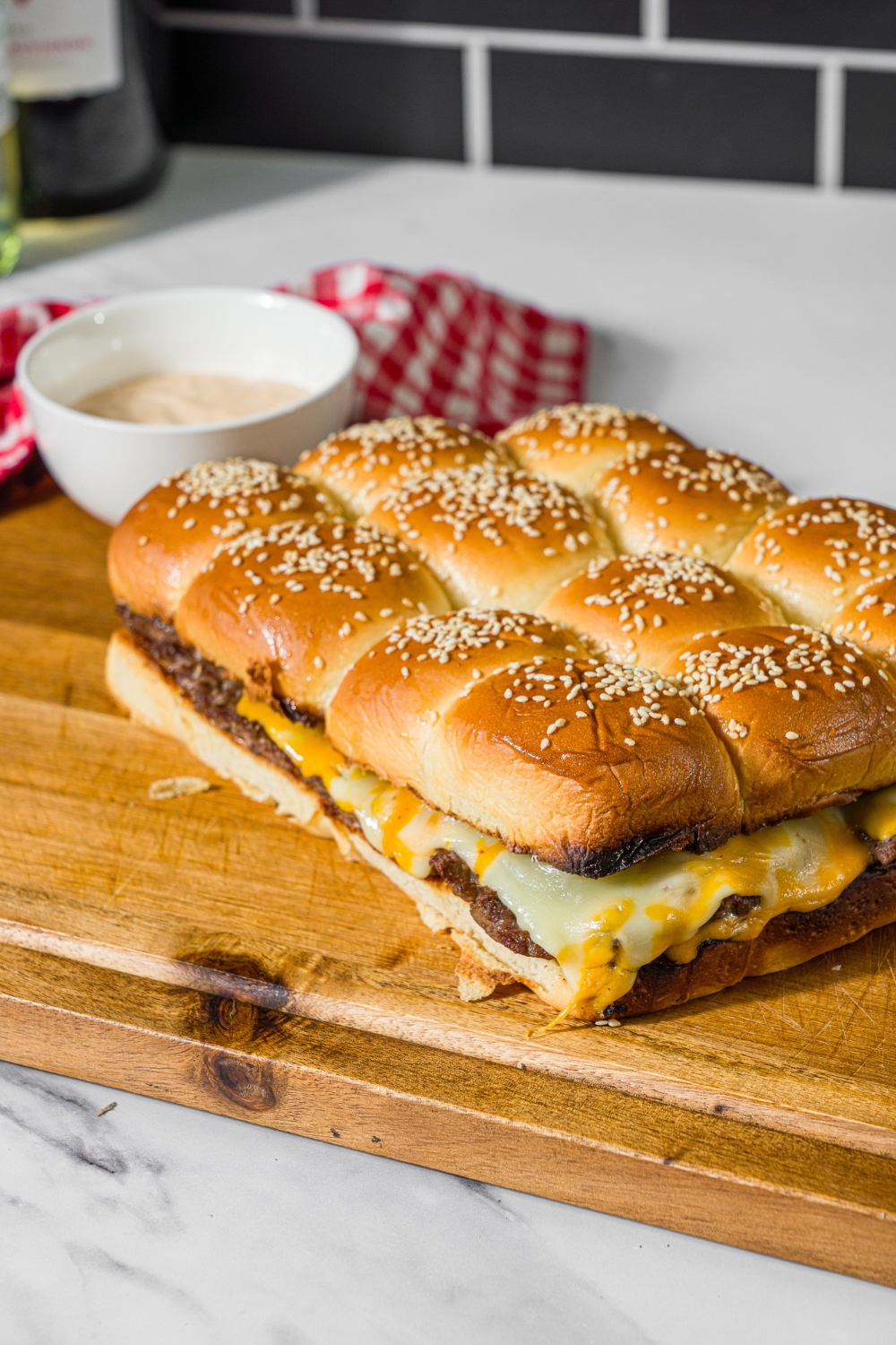 A wooden board with a set of cheeseburger sliders on hawaiian rolls. The board is on a marble counter with a bowl of burger sauce and red checkered napkin.