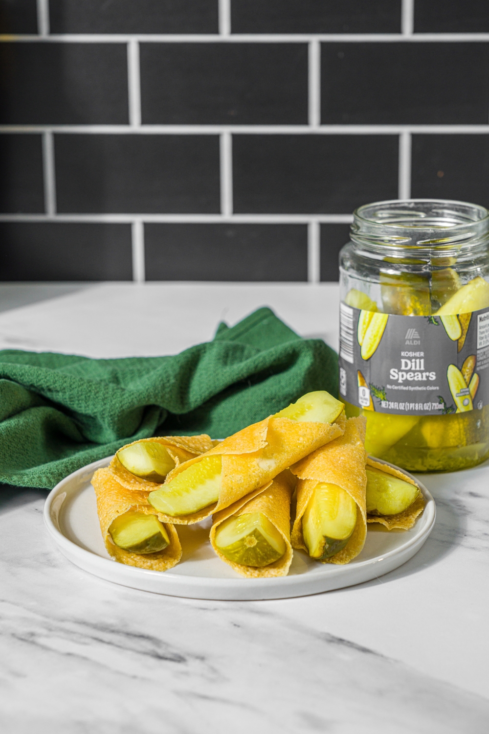 A plate with a pile of chickles made with pickles and cheese. The plate is on a marble counter with a green cloth napkin and a jar of pickles.