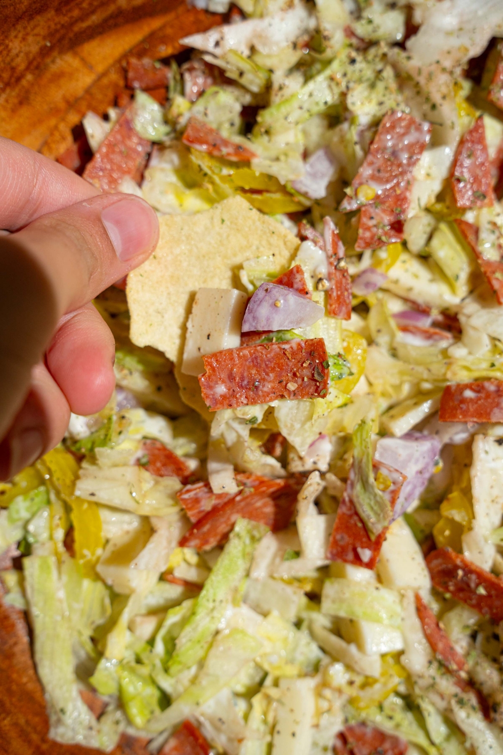A large wooden bowl with Italian grinder dip including chopped lettuce, salami, pepperoni, red onion, banana peppers, and cheese. There is a chip being dipped into the bowl.