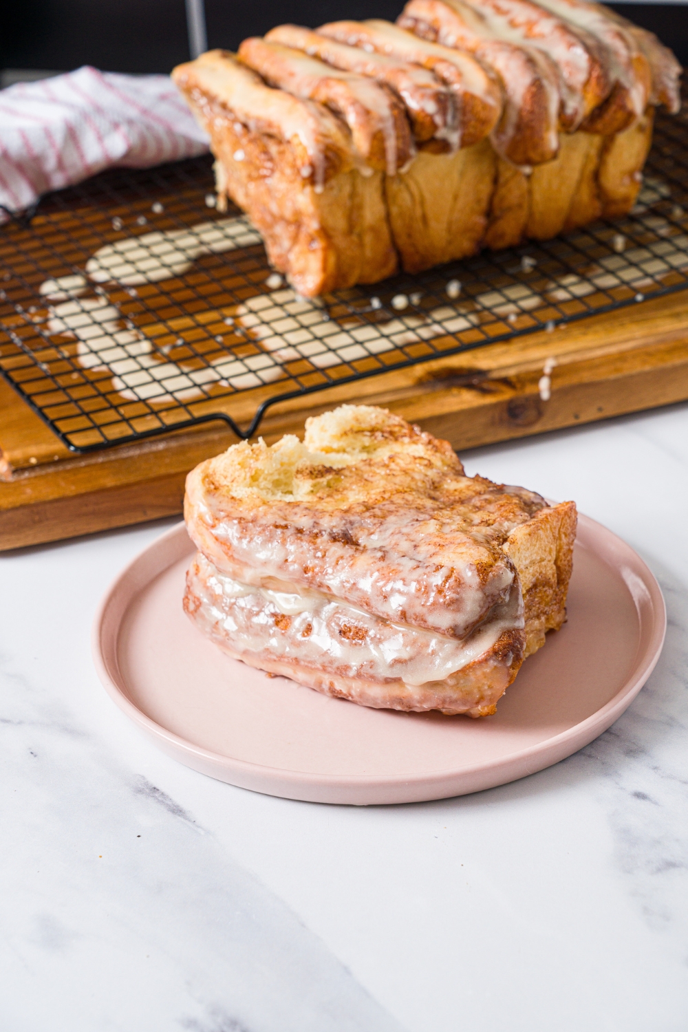 A pink plate with a slice of cinnamon pull apart bread topped with glaze. The plate is on a marble counter with a loaf of bread on a wooden board with a wire rack.