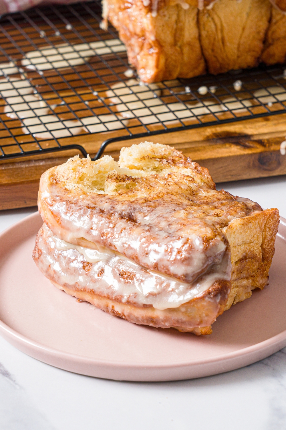A pink plate with a slice of cinnamon pull apart bread topped with glaze. The plate is on a marble counter with a loaf of bread on a wooden board with a wire rack.