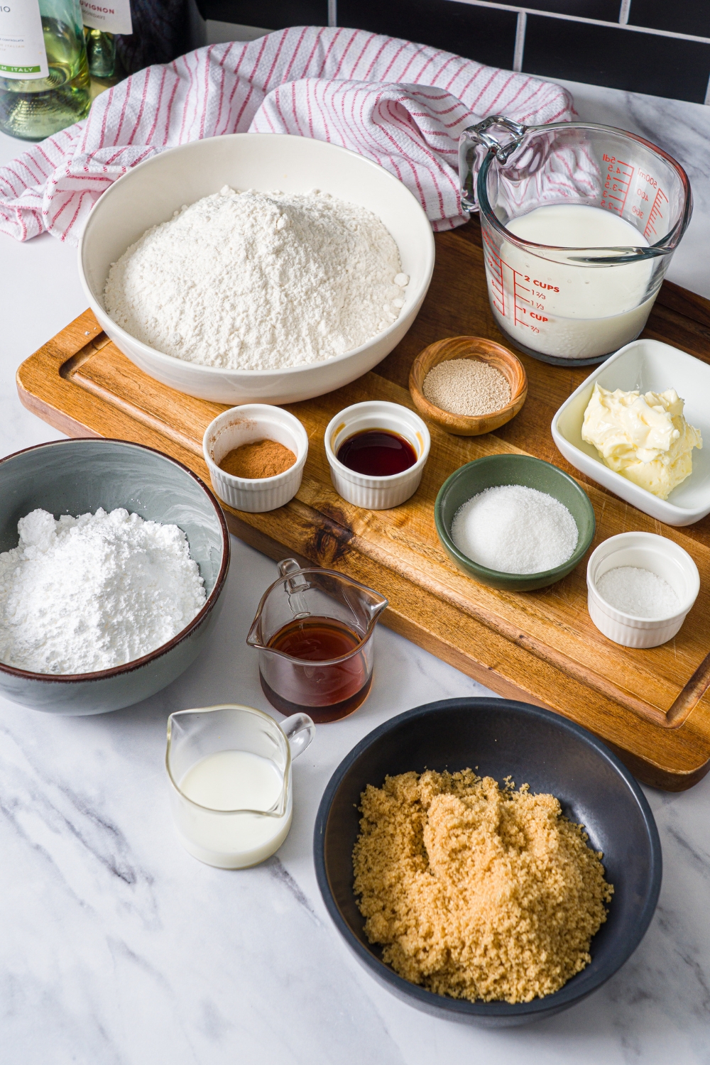 A marble counter with a wooden board with ingredients to make cinnamon pull apart bread including flour, yeast, milk, sugar, egg, vanilla, brown sugar, cinnamon, powdered sugar, and butter.