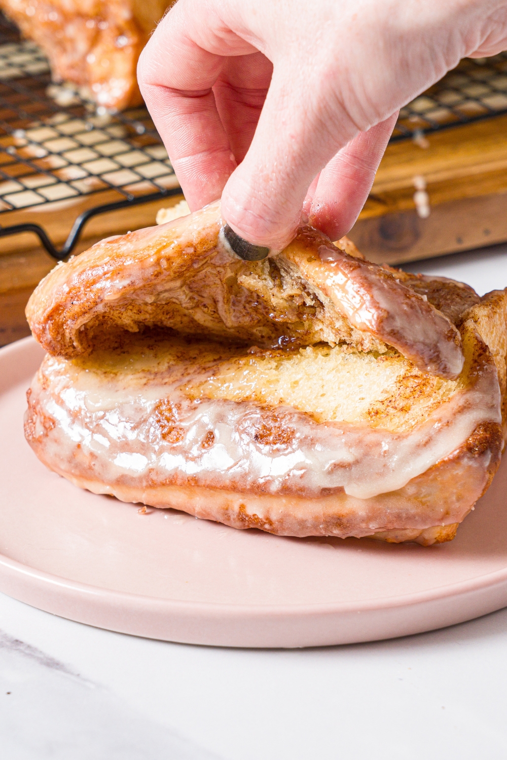 A pink plate with a slice of cinnamon pull apart bread topped with glaze. A hand is pulling apart the top layer. The plate is on a marble counter with a loaf of bread on a wooden board with a wire rack.
