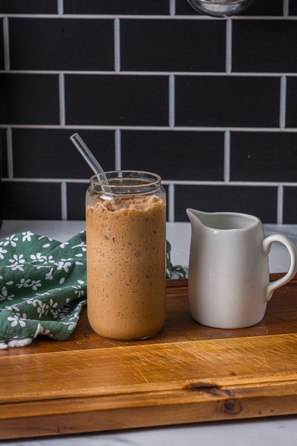 A wooden board with a glass of coffee slushie with a straw in the glass. There is a carafe of milk on the board with a green cloth napkin.