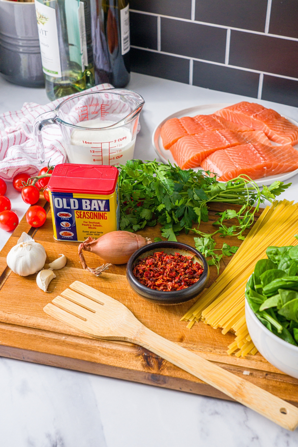 A wooden board with ingredients to make tuscan salmon with pasta including salmon, spinach, linguine, shallot, garlic, fresh parsley, old bay, tomatoes, heavy cream, and spinach.
