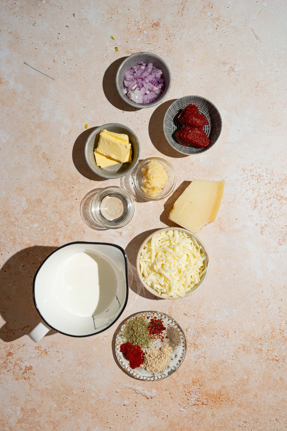 A tan counter with several bowls containing ingredients to make creamy tomato pasta sauce including garlic, onion, tomato paste, heavy cream, butter, shredded cheese, and seasonings.
