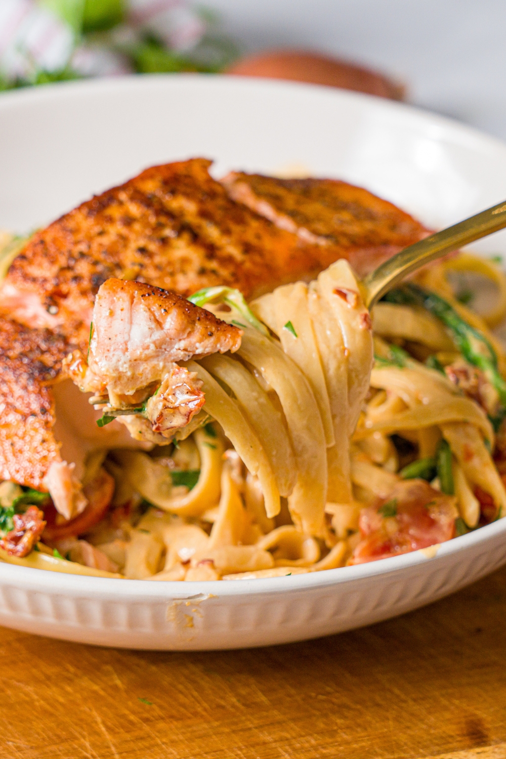 A white bowl with tuscan salmon with pasta. A fork is pulling a bite of pasta from the bowl. The bowl is on a wooden board.