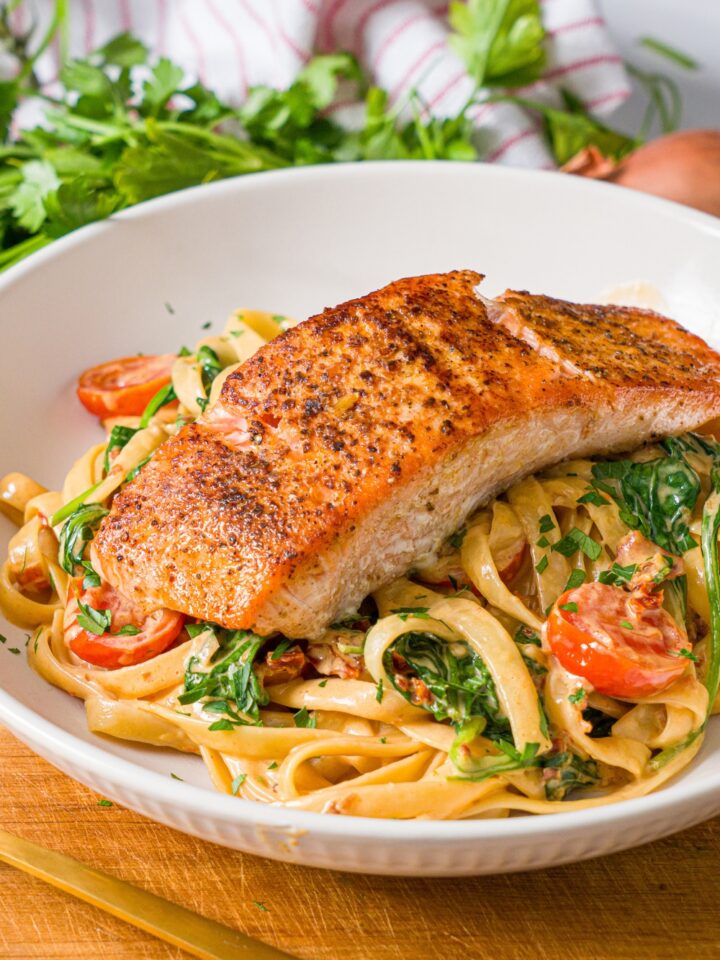 A white bowl with creamy tuscan pasta with seared salmon on top. The bowl is on a wooden board with a fork. The board is on a marble counter with fresh parsley.