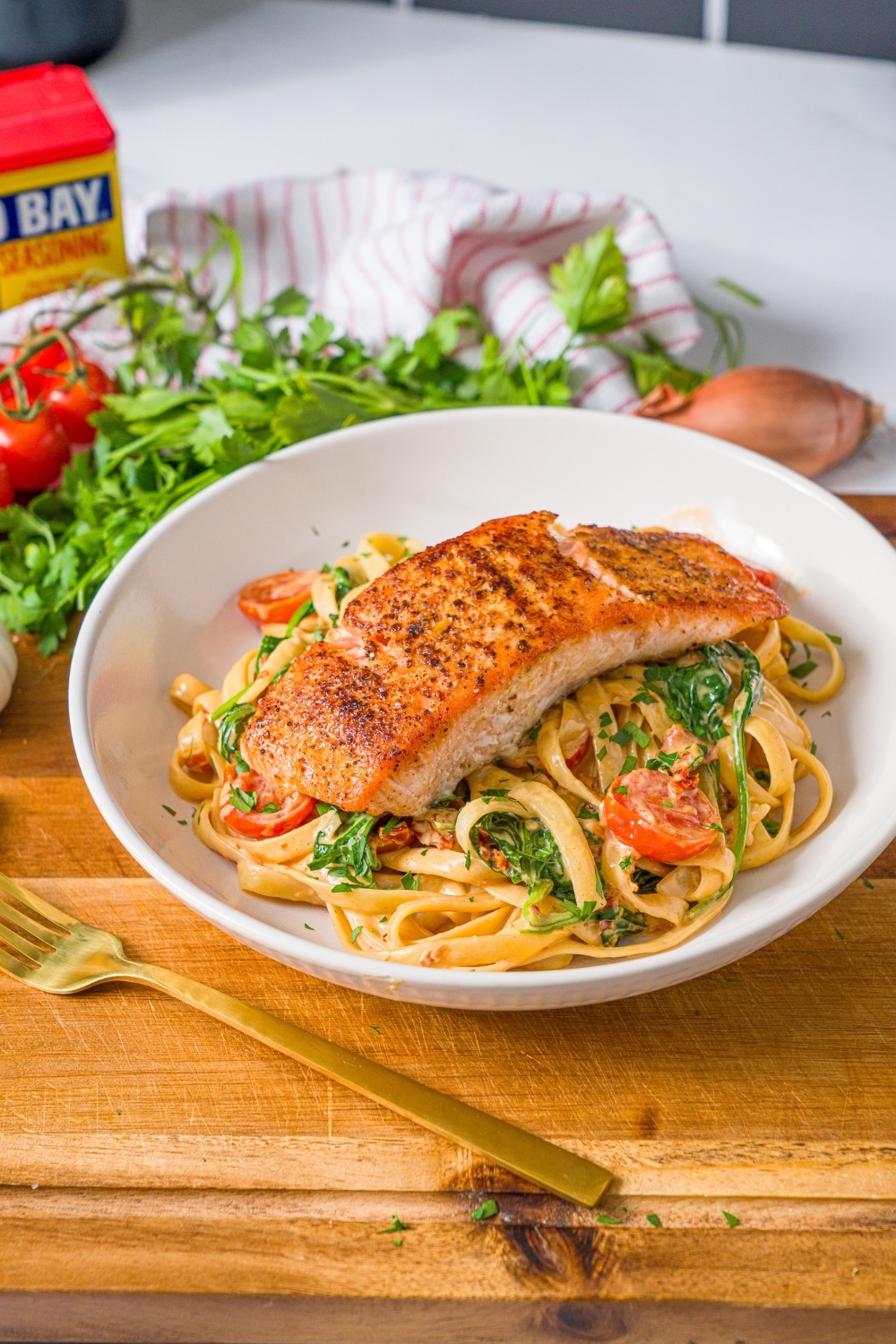 A white bowl with creamy tuscan pasta with seared salmon on top. The bowl is on a wooden board with a fork. The board is on a marble counter with fresh parsley.