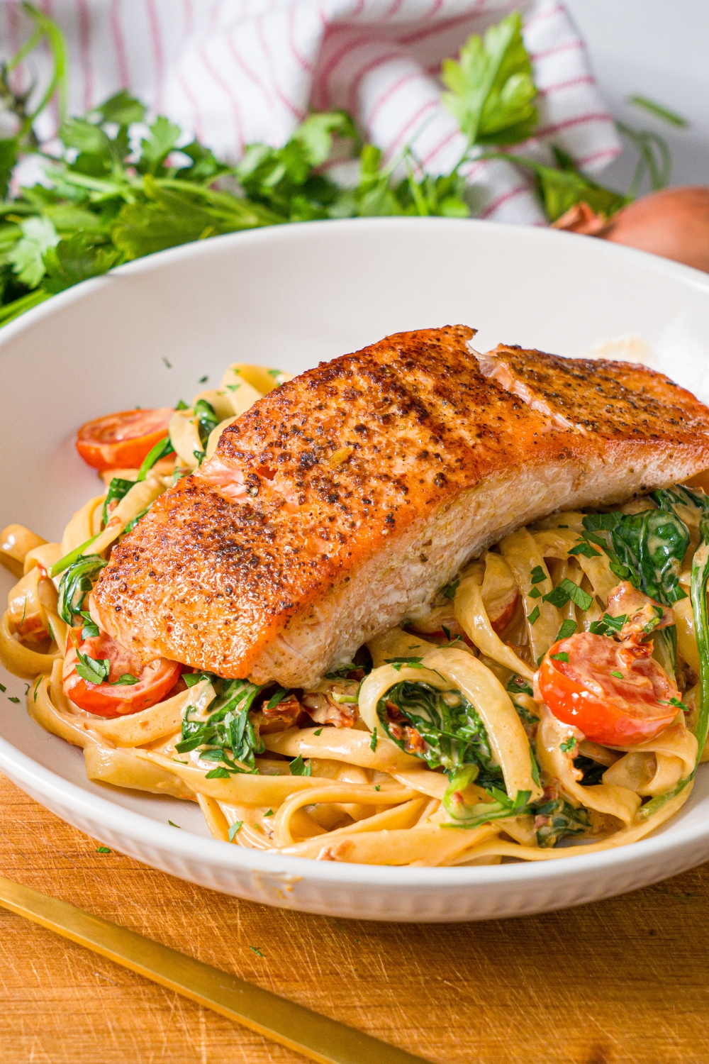A white bowl with creamy tuscan pasta with seared salmon on top. The bowl is on a wooden board with a fork. The board is on a marble counter with fresh parsley.