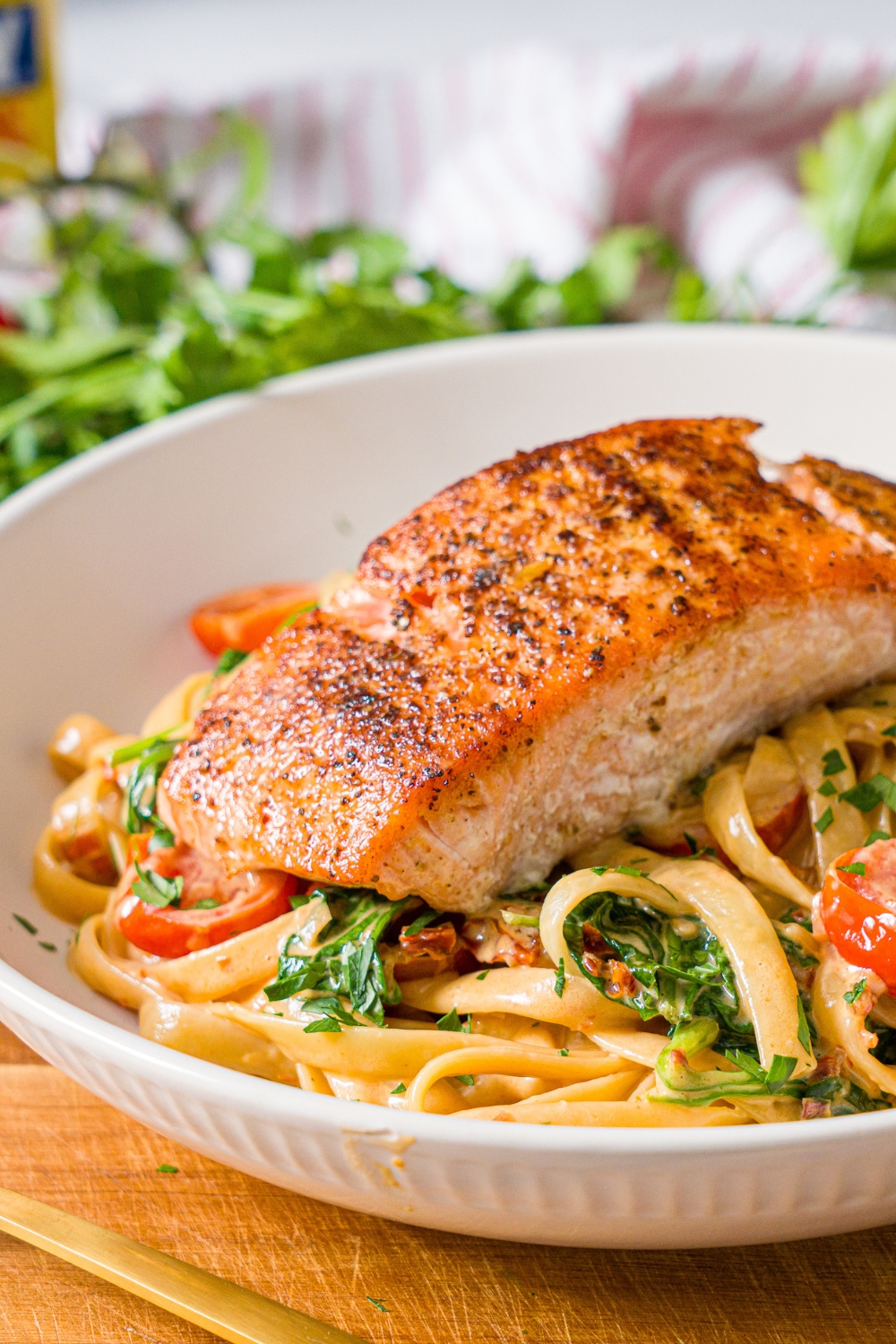 A white bowl with creamy tuscan pasta with seared salmon on top. The bowl is on a wooden board. The board is on a marble counter with fresh parsley.