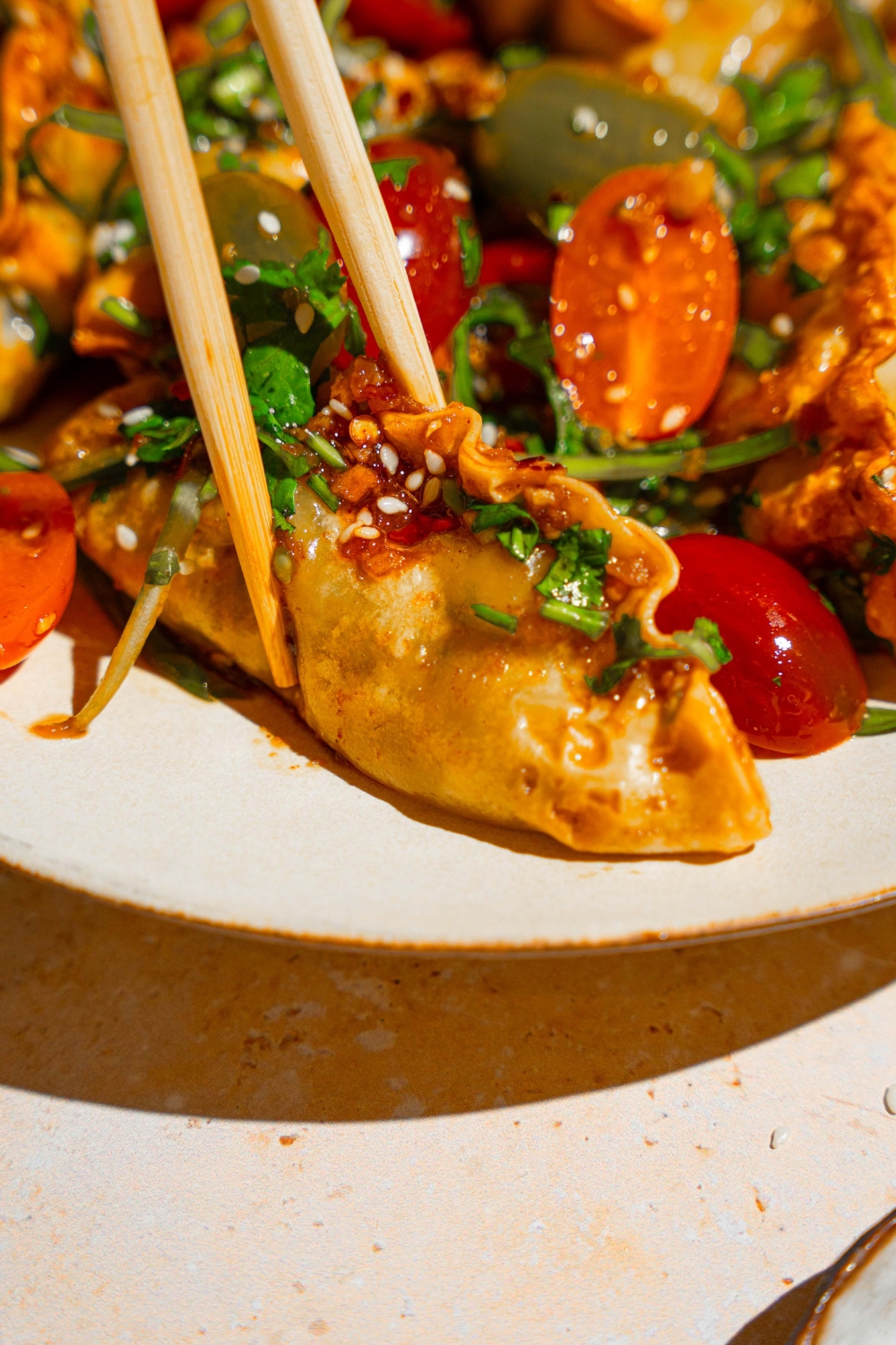 A white plate with crispy dumpling salad with crispy dumplings, sliced tomatoes, and scallions, tossed in a soy sauce and chili oil dressing. A pair of chopsticks are taking a dumpling from the salad.