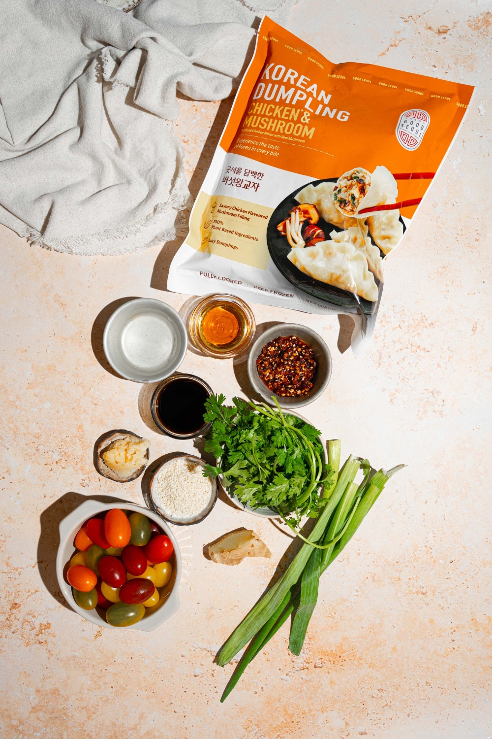 A tan counter with several bowls containing ingredients to make crispy dumpling salad including frozen dumplings, tomatoes, scallions, cilantro, soy sauce, chili oil, garlic, ginger, and more.