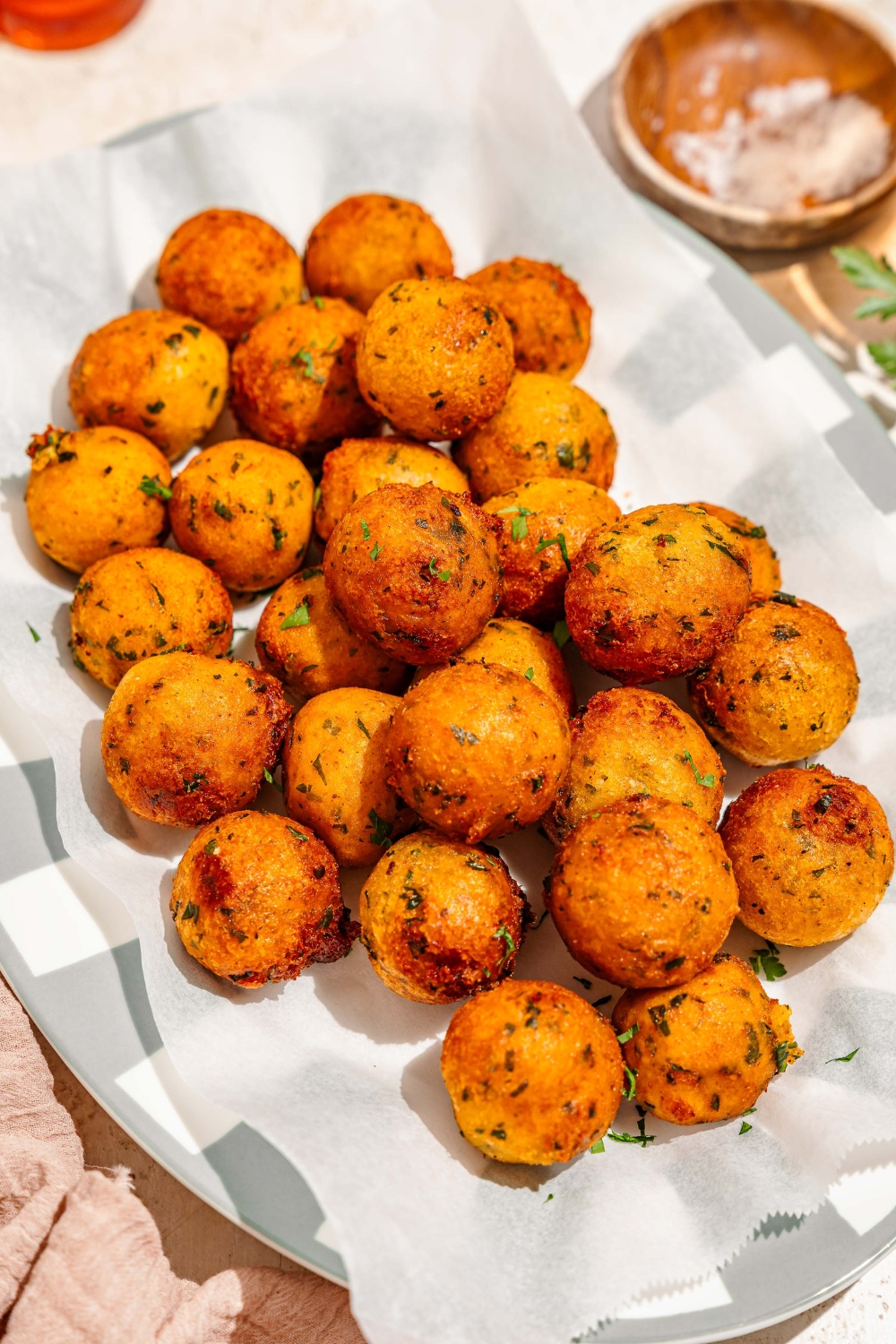 A platter lined with parchment paper with crispy potato balls garnished with fresh parsley. The platter is on a tan counter with a small bowl of salt.