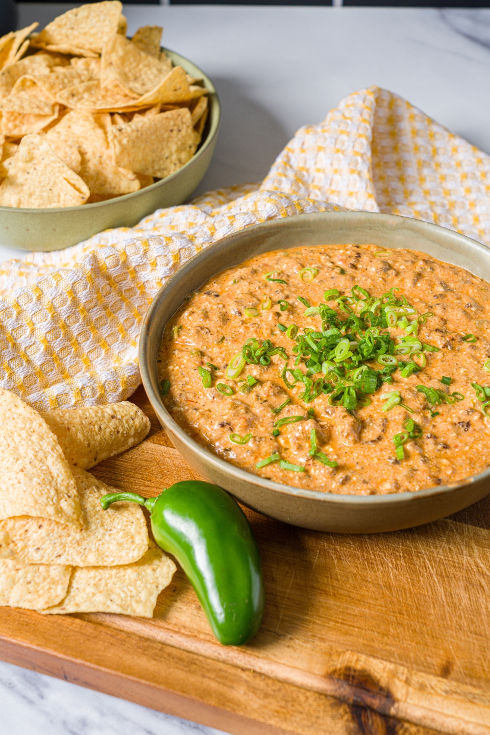 A bowl of divorced dad dip garnished with sliced green onion on a wooden board with a jalapeño. The board is on a white counter with a bowl of tortilla chips.
