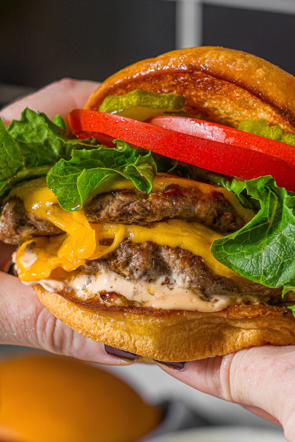 Hands holding a double smash burger with lettuce, tomato, caramelized onion, burger sauce, and pickles.