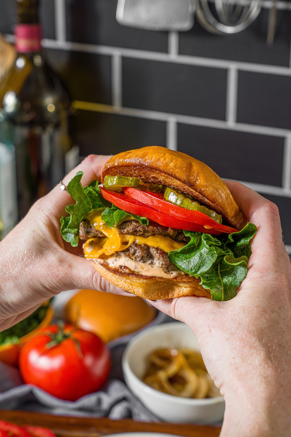 A close up of hands holding a double smash burger with lettuce, tomato, caramelized onion, burger sauce, and pickles. There is a counter with bowls of ingredients blurred in the background.