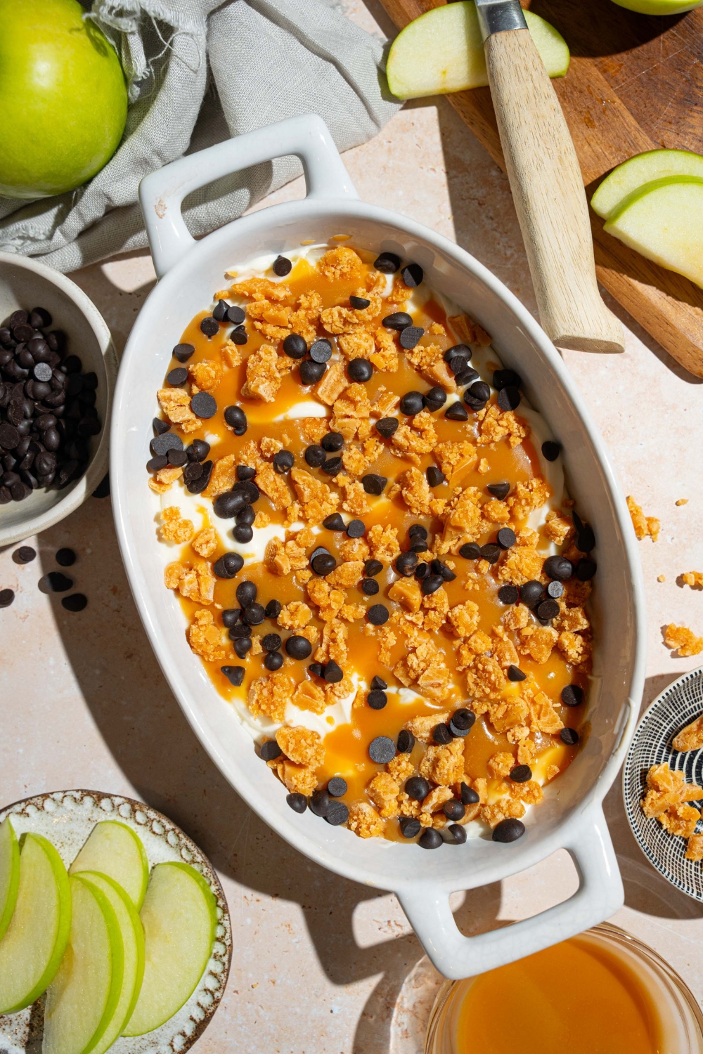 An oval platter with caramel apple cheesecake dip topped with toffee bits and chocolate chips. The platter is on a tan counter with a plate of sliced apples and additional ingredients.