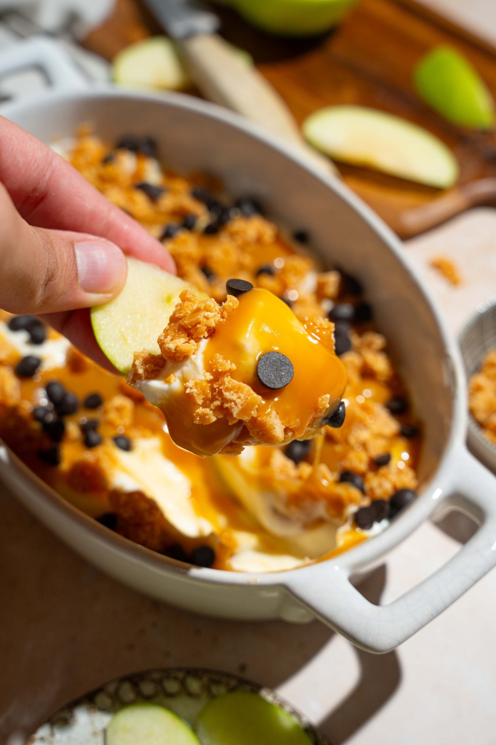 A close up of a hand holding an apple slice dipped in caramel apple cheesecake dip topped with toffee bits and chocolate chips. There is a platter of cheesecake dip blurred in the background.