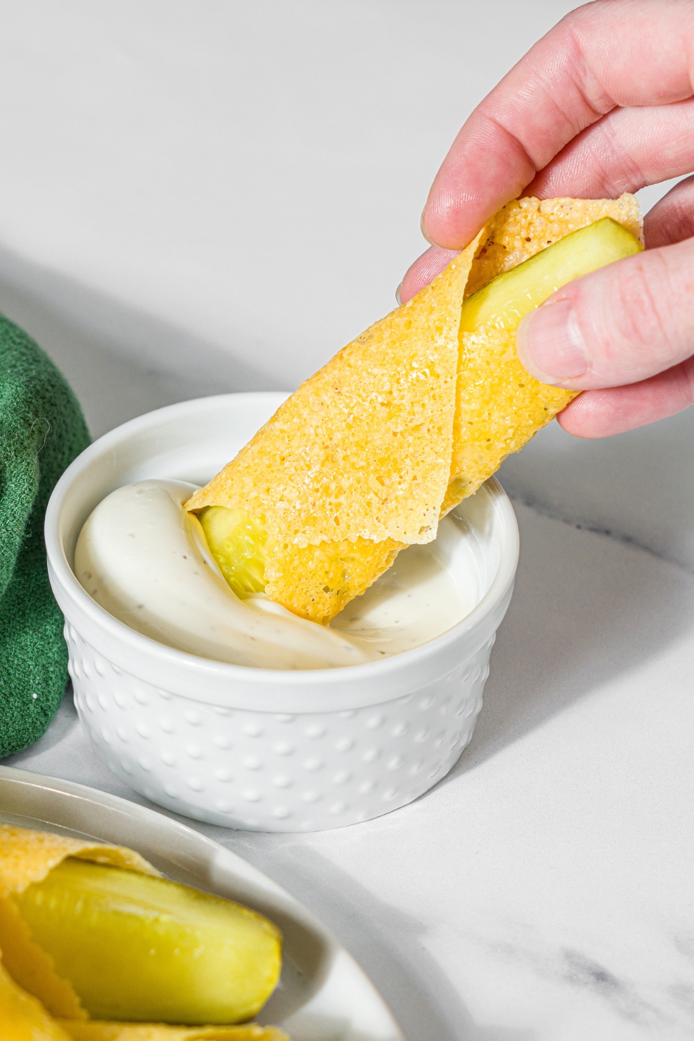 A hand dipping a chickle into a small bowl of dip. The dip is on a marble counter with a plate of chickles.