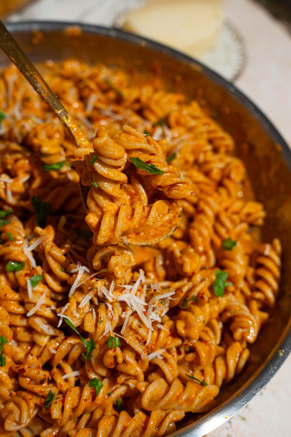 A skillet with fusilli pasta tossed in creamy tomato pasta sauce garnished with fresh basil and shredded cheese. A fork is taking a bite from the skillet. The skillet is on a tan counter with small bowls of garnishes and a pepper mill.