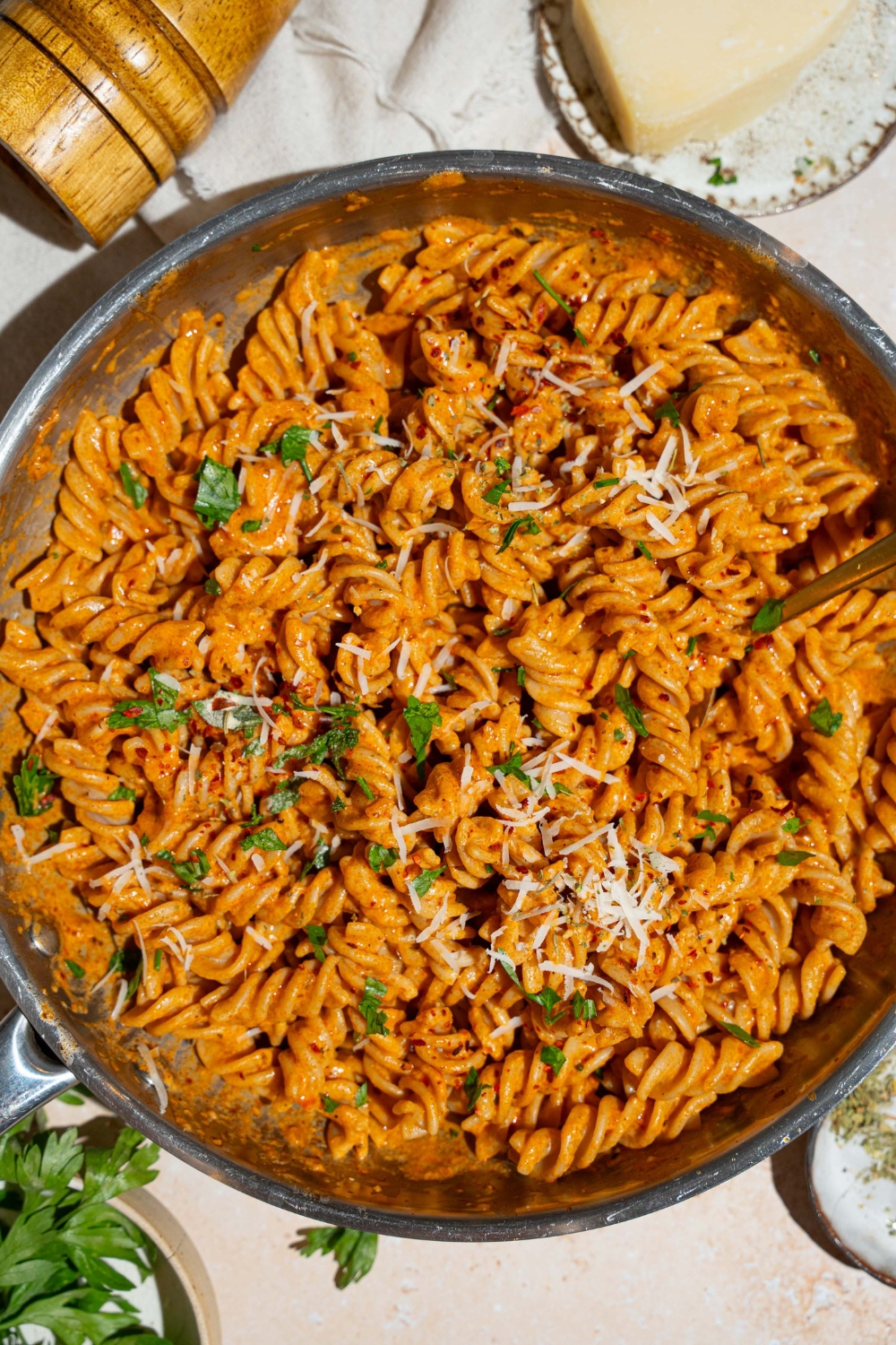 A skillet with fusilli pasta tossed in creamy tomato pasta sauce garnished with fresh basil and shredded cheese. There is a fork in the pasta. The skillet is on a tan counter with small bowls of garnishes and a pepper mill.