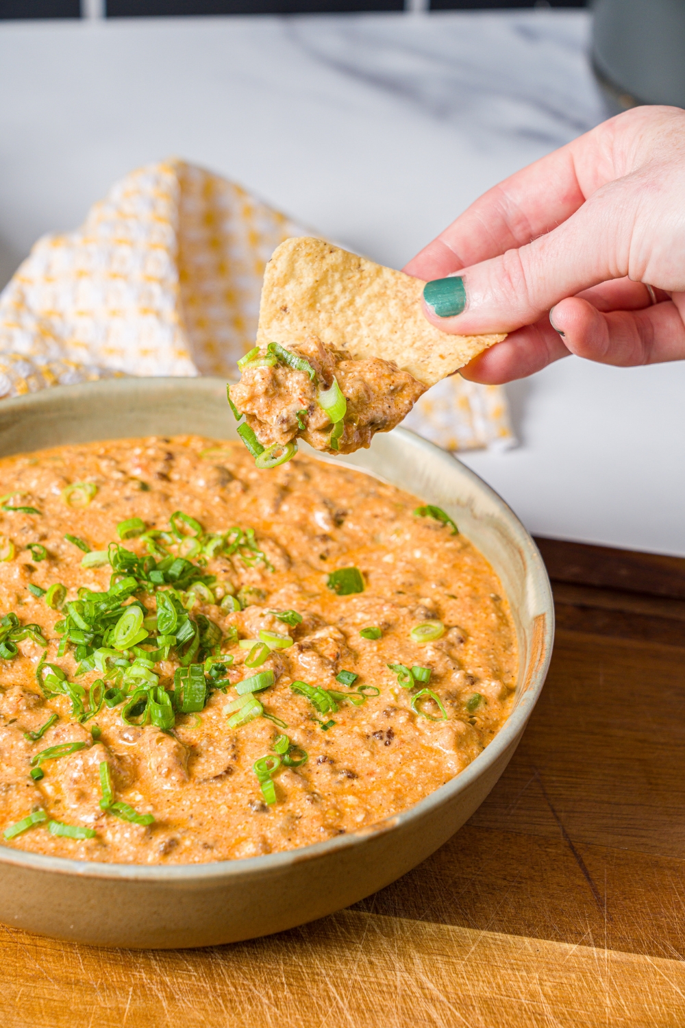 A bowl of divorced dad dip garnished with sliced green onion on a wooden board. A hand is holding a chip that was dipped in the bowl. The board is on a white counter with a bowl of tortilla chips.