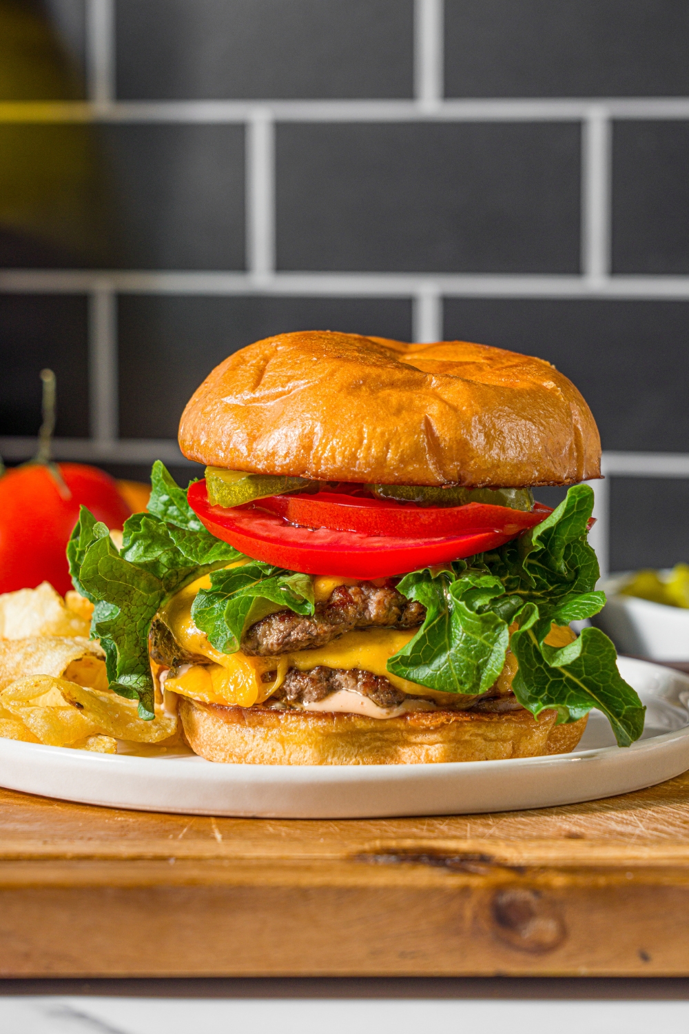 A white plate with a double smash burger with lettuce, caramelized onions, tomato, pickles, and burger sauce served with chips. The plate is on a wooden board.
