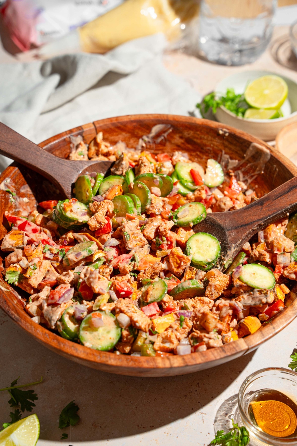 A large wooden bowl with hot honey chicken bowl including sweet potato, chicken, cucumbers, onion, and bell pepper tossed in a hot honey sauce. There are two salad spoons in the bowl. The bowl is on a tan counter with small bowls of ingredients.