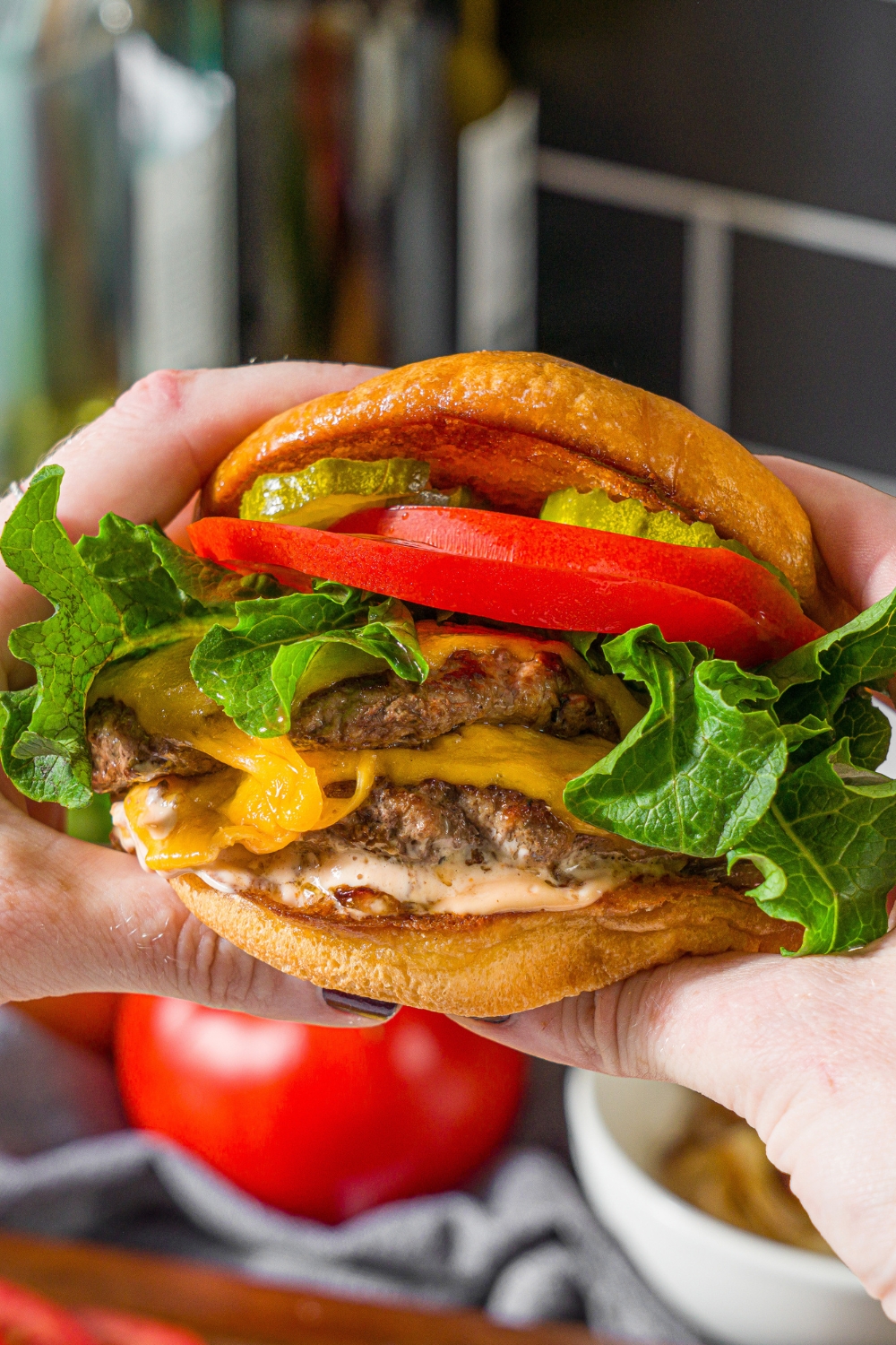 A close up of hands holding a double smash burger with lettuce, tomato, caramelized onion, burger sauce, and pickles. There is a counter with bowls of ingredients blurred in the background.