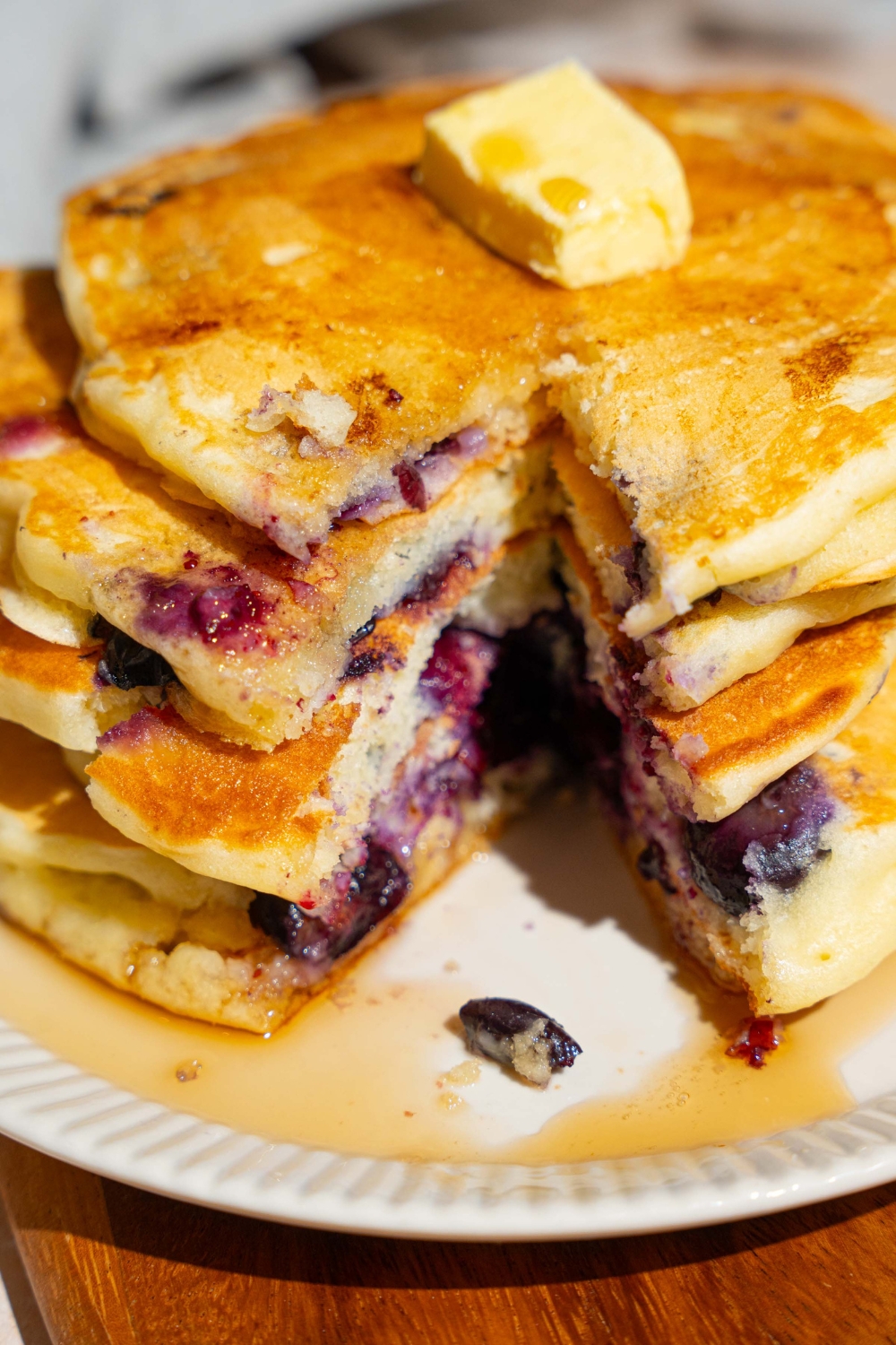 A white plate with a stack of buttermilk blueberry pancakes topped with butter and drizzled with syrup. There is a bite taken from the stack. The plate is on a wooden board.