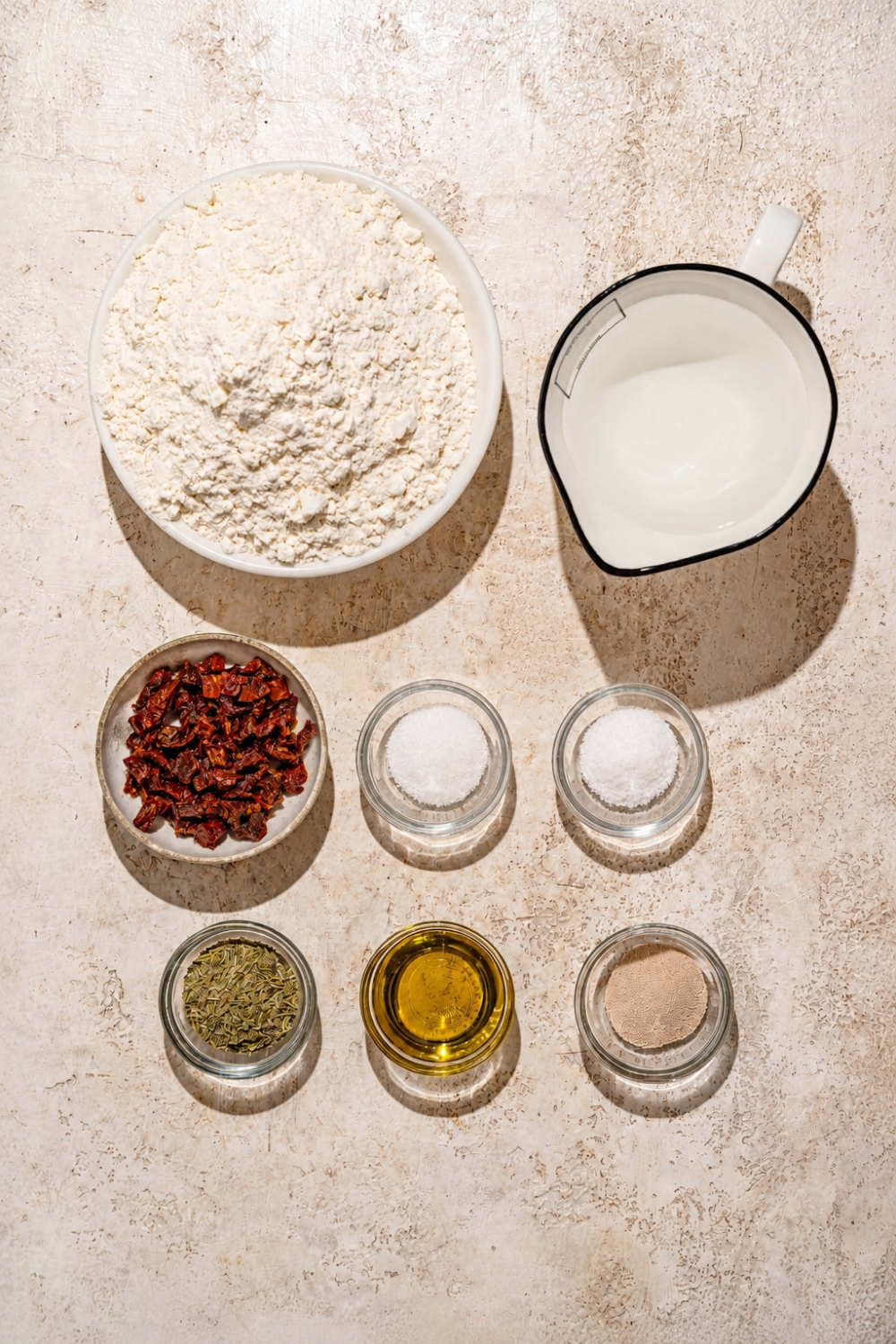 A tan counter with several bowls containing ingredients to make focaccia muffins including flour, yeast, water, sugar, salt, olive oil, and rosemary