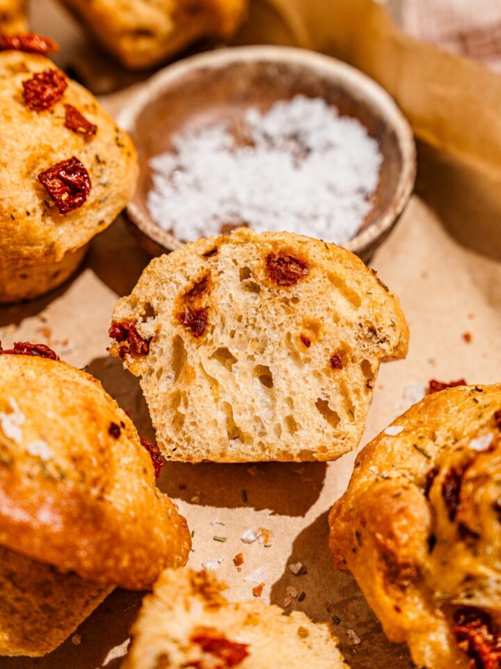A baking pan lined with parchment paper with rosemary focaccia muffins and small bowl of salt. One of the muffins is sliced in half.