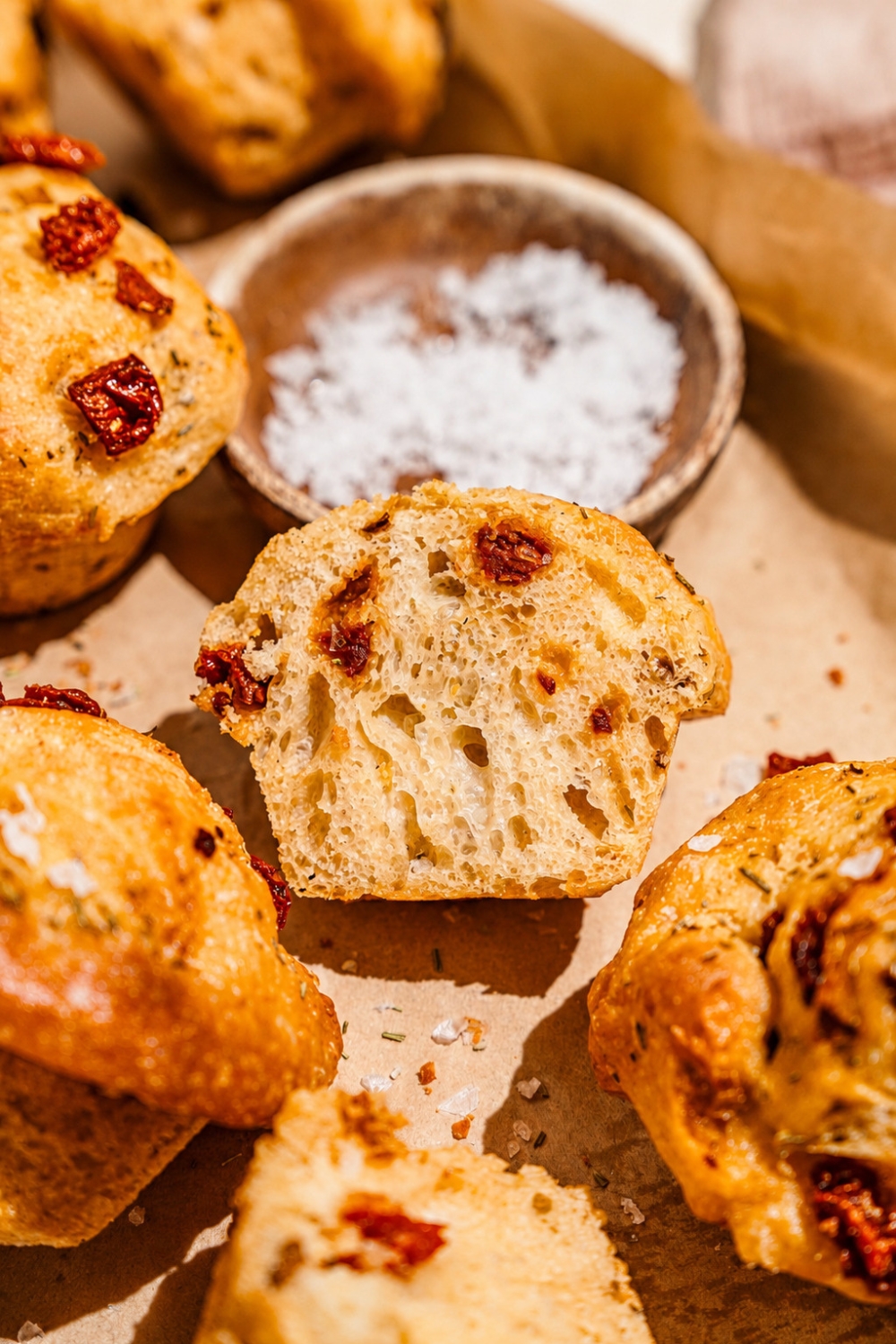 A baking pan lined with parchment paper with rosemary focaccia muffins and small bowl of salt. One of the muffins is sliced in half.