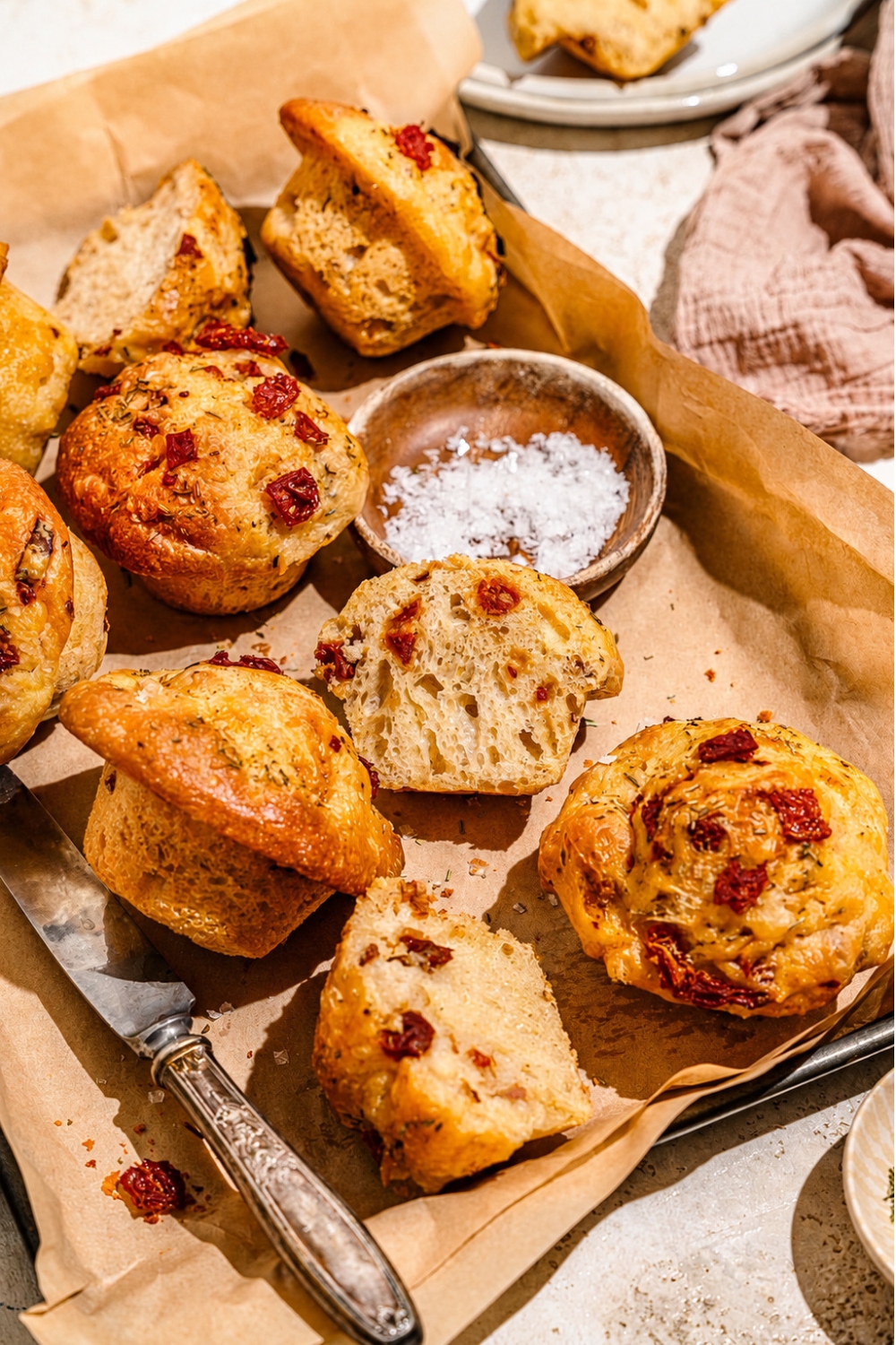 A baking pan lined with parchment paper with rosemary focaccia muffins with a knife and small bowl of salt. One of the muffins is sliced in half. The pan is on a tan counter.