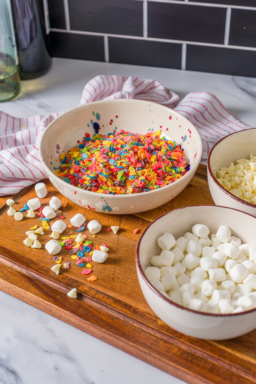 A wooden board with ingredients to make fruity pebbles rice crispy treats including fruity pebbles, marshmallows, and white chocolate chips.