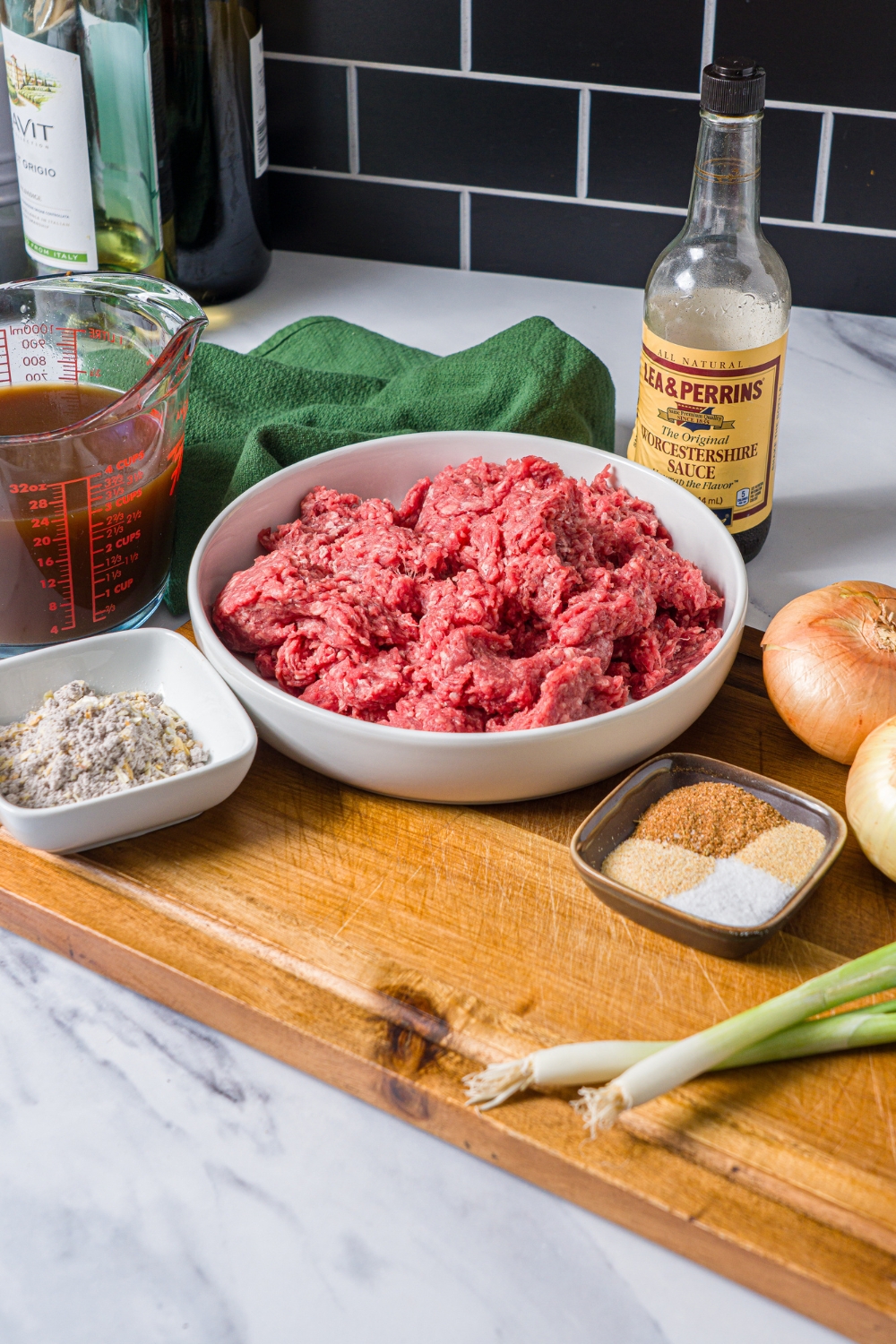 A wooden board with ingredients to make hamburger steak with onion gravy including ground beef, Worcestershire sauce, onions, green onion, beef broth, and seasonings.