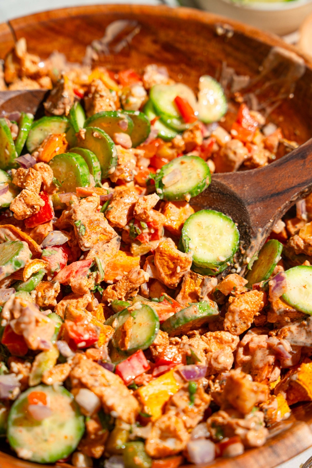A large wooden bowl with hot honey chicken bowl including sweet potato, chicken, cucumbers, onion, and bell pepper tossed in a hot honey sauce. A salad spoon is scooping the mixture.