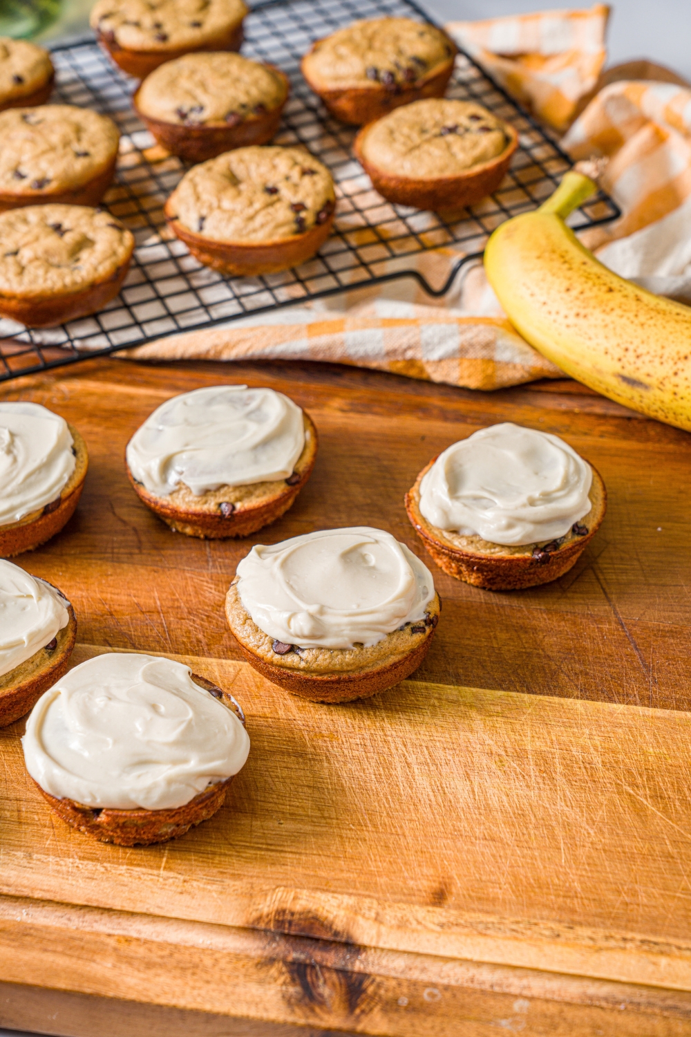 A wooden board with frosted protein banana muffins. The board is on a marble counter with a wire rack of muffins and a banana.