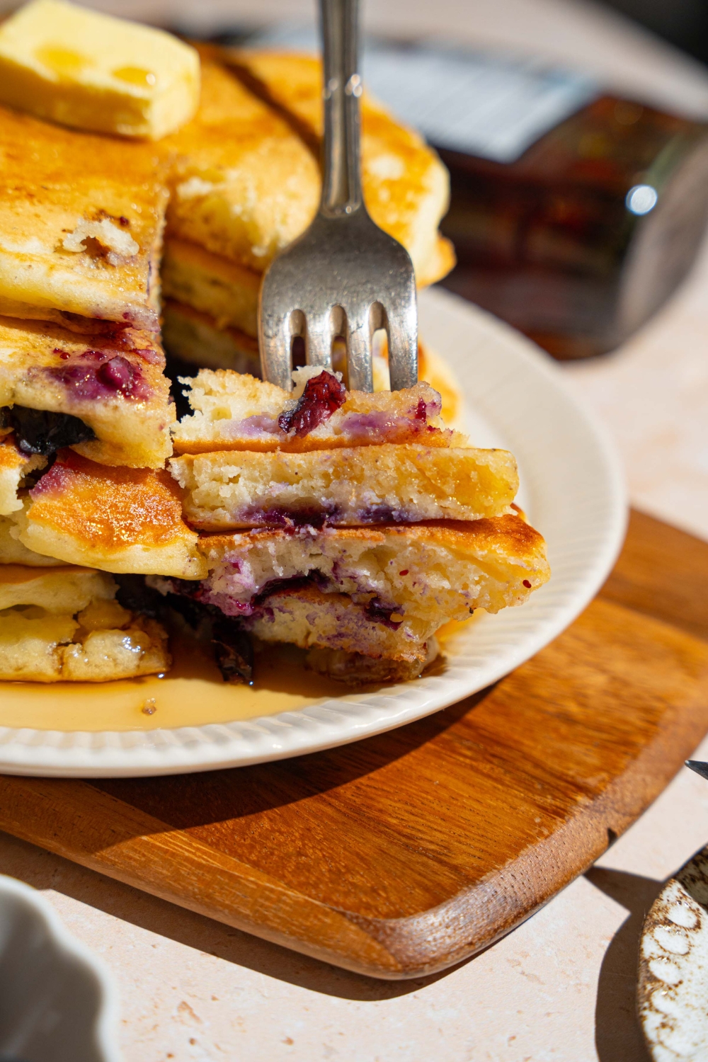 A white plate with a stack of buttermilk blueberry pancakes. A fork is taking a bite from the stack. The plate is on a wooden board.
