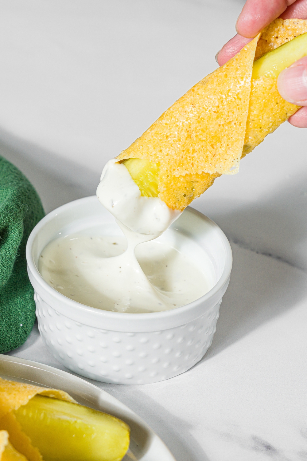 A hand dipping a chickle into a small bowl of dip. The dip is on a marble counter with a plate of chickles.