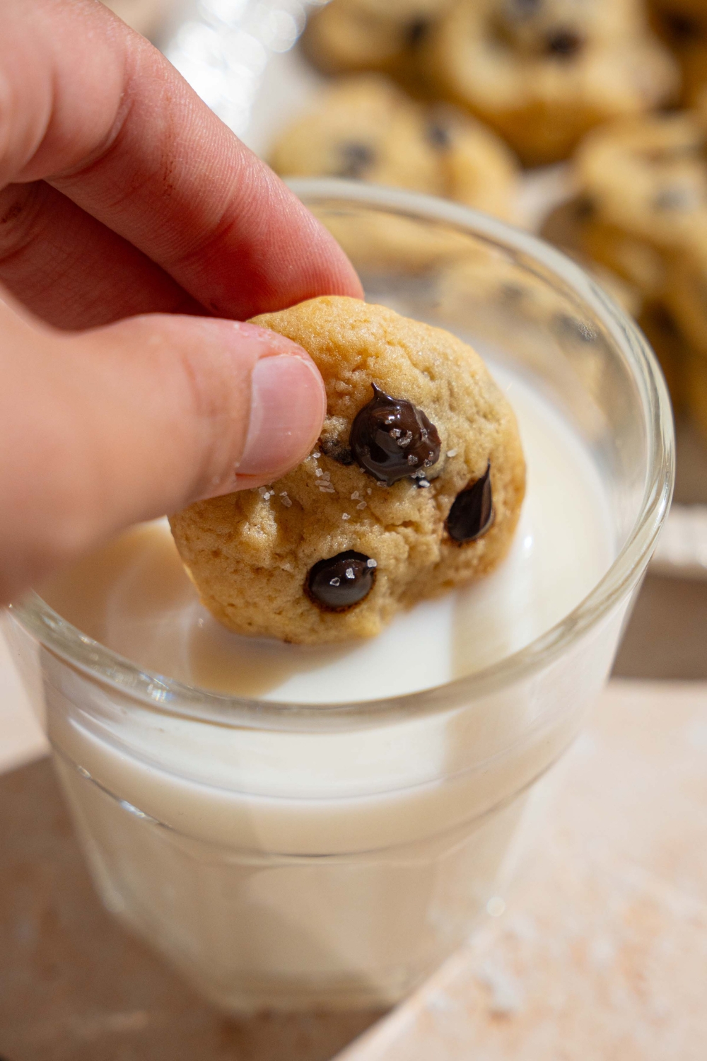 A hand dipping a mini chocolate chip cookie into a glass of milk. The glass is on a tan counter with a plate of cookies.