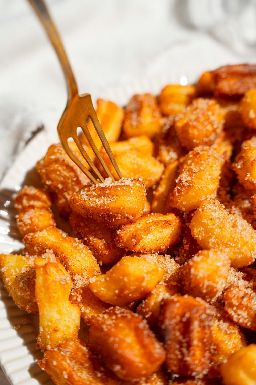 A white plate with mini churros coated in cinnamon sugar. A fork is taking a churro from the plate. The plate is on a tan counter.