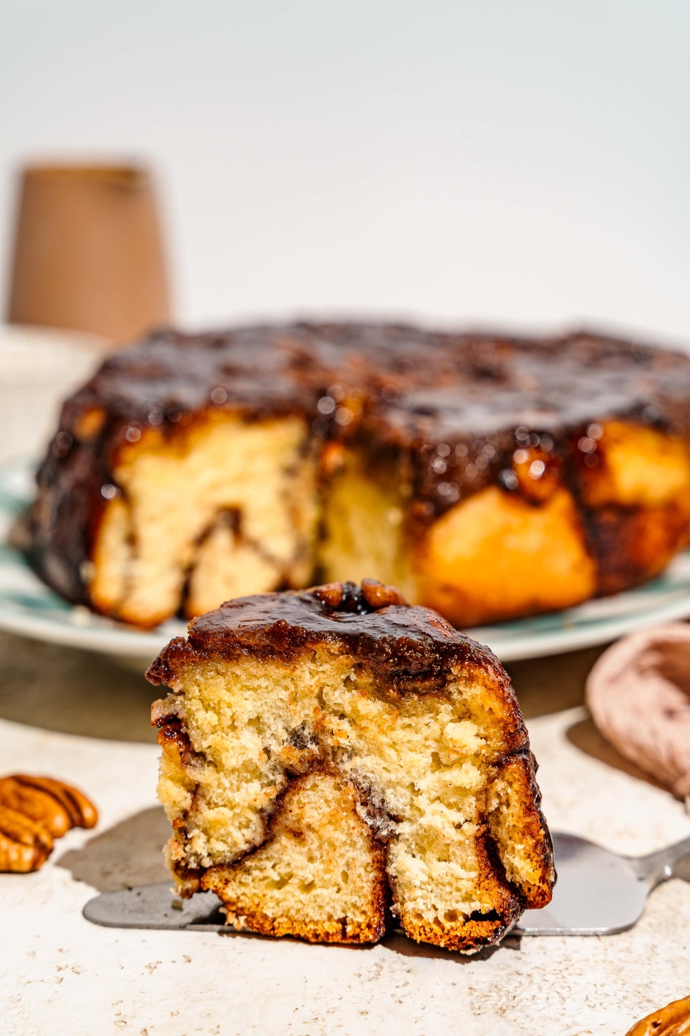 A close up of a white plate with a pecan caramel sticky bun and a fork. There is a tray of sticky buns on a tan counter blurred in the background.