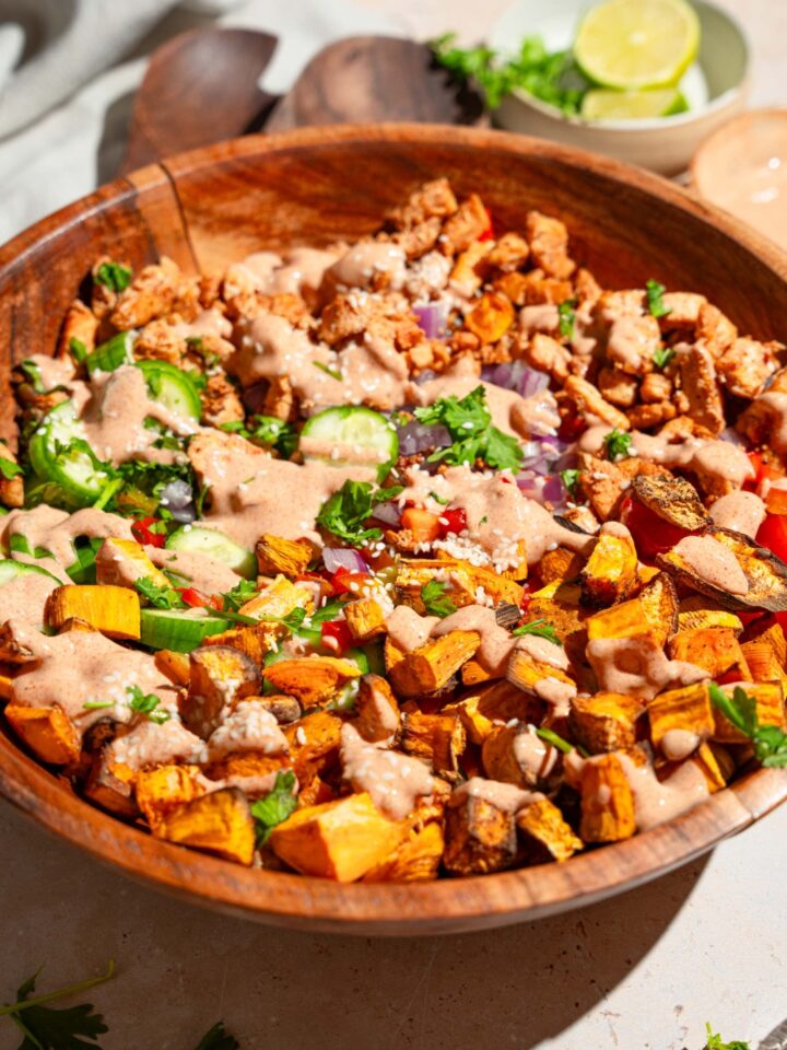 A large wooden bowl with hot honey chicken bowl including sweet potato, chicken, cucumber, onion, and bell peppers drizzled with hot honey sauce. The bowl is on a tan counter with small bowls of ingredients.