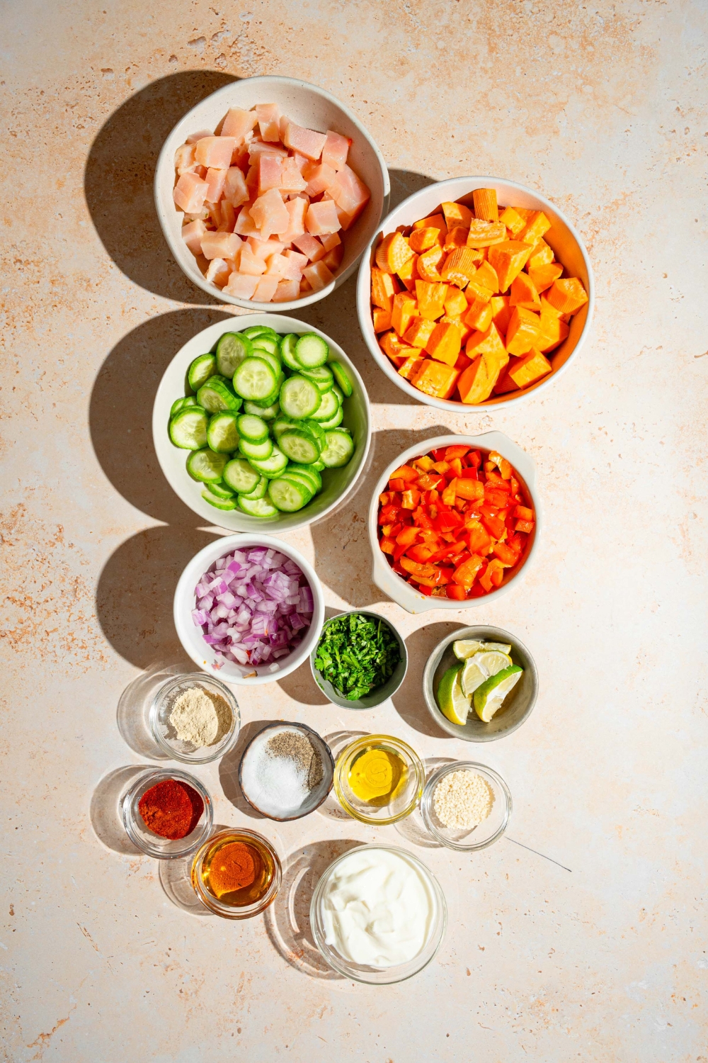 A tan counter with several bowls containing ingredients to make hot honey chicken bowls including chicken, sweet potato, cucumbers, bell peppers, lime, red onion, cilantro, greek yogurt, honey, and seasonings.