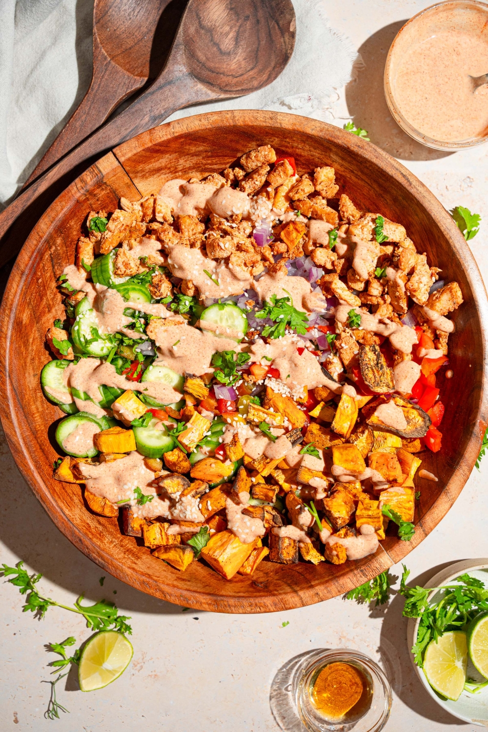 A large wooden bowl with hot honey chicken bowl including sweet potato, chicken, cucumber, onion, and bell peppers drizzled with hot honey sauce. The bowl is on a tan counter with small bowls of ingredients.